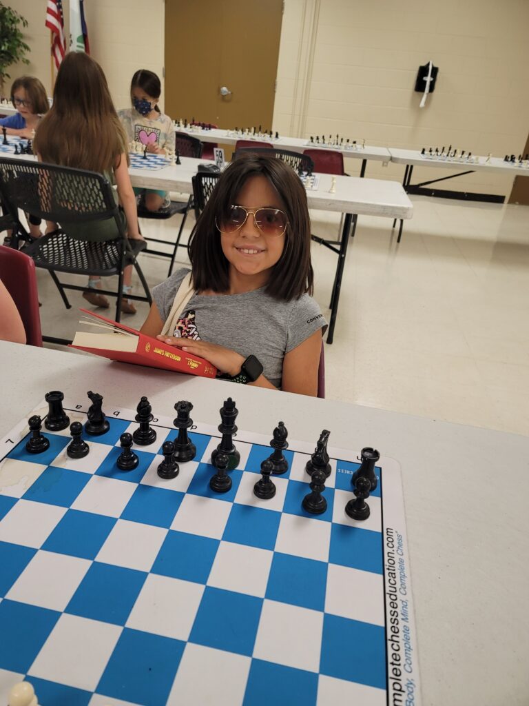 young girl with sunglasses holding a book while playing chess